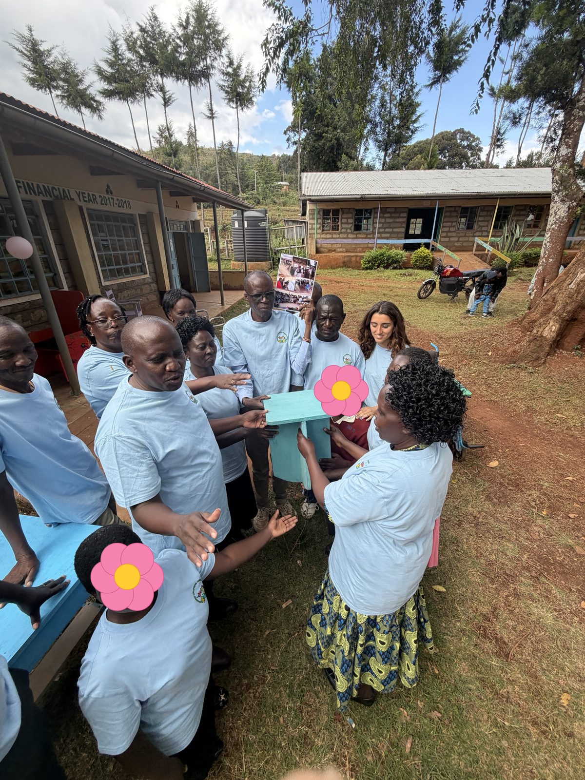 Teachers in workshop T-shirts gathered around a turquoise wooden stool outside the school, holding it together with learners while one teacher speaks; learners' faces are covered with flowers for privacy