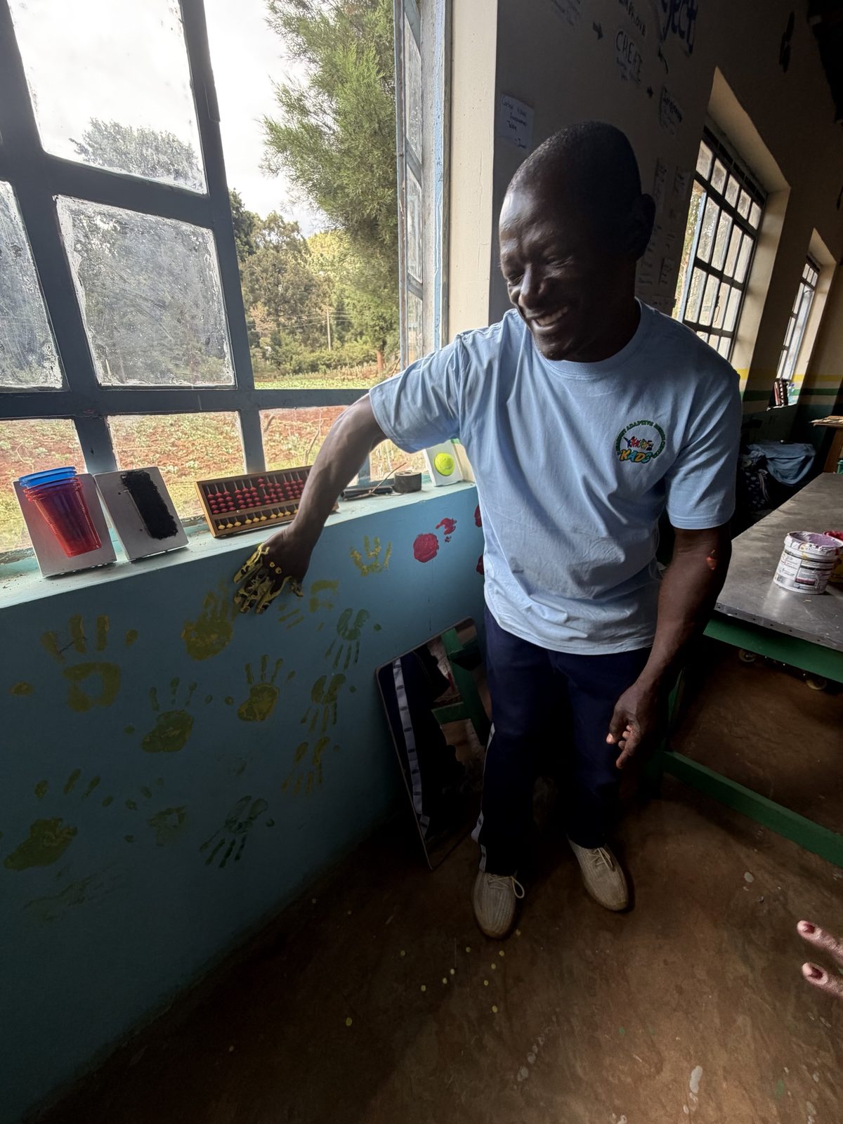 A teacher laughing as he presses a yellow-painted hand onto the wall under the workshop window, among dozens of yellow and red handprints, with tactile cue cards on the sill