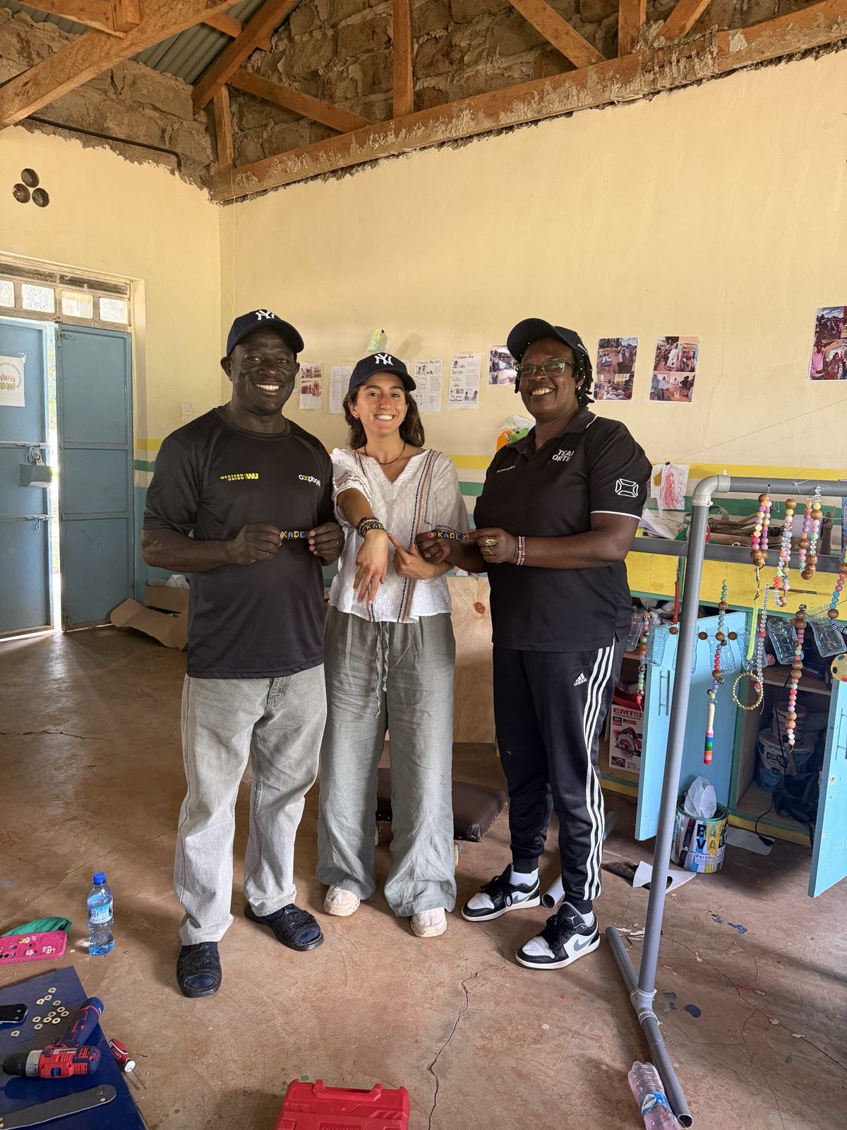 Leilani standing in the workshop between the two teachers taking over the workshop, all in matching navy caps and holding up beaded bracelets that spell KADC
