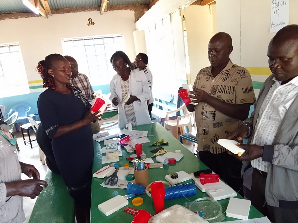 Teachers at a long green workbench covered in red and blue cups, bottle caps and white boards, each holding a tactile cue card they are assembling