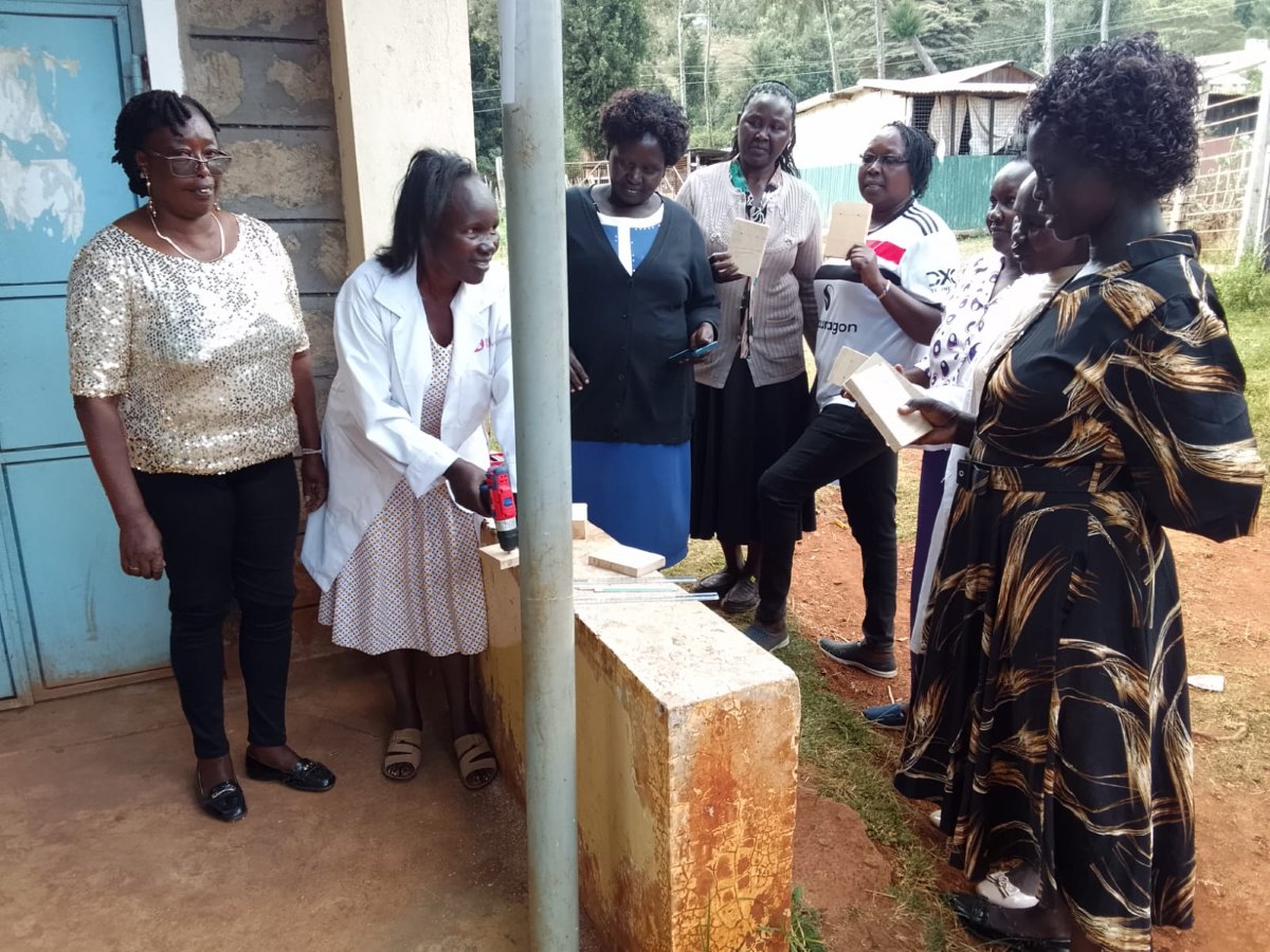 A teacher in a white coat drilling into a wooden board on a low wall outdoors while a group of women teachers holding their own boards look on