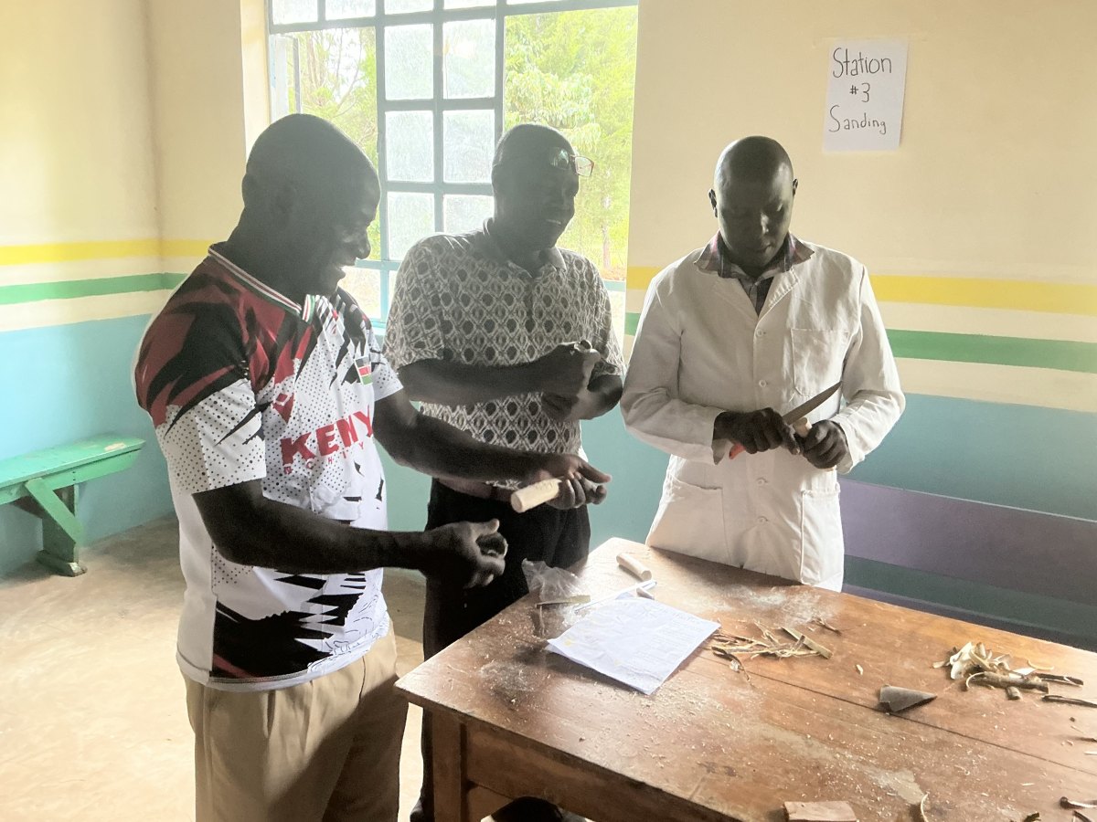 Three teachers at the sanding station, laughing as they carve and sand wooden spoon handles with a knife and sandpaper