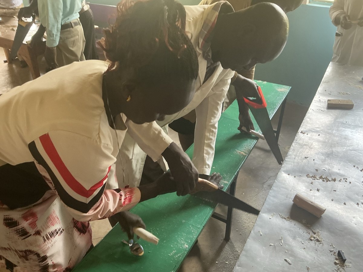 Two teachers in white coats bent over a green bench, sawing wooden blocks to length for spoon handles, with a spoon and a finished handle beside them
