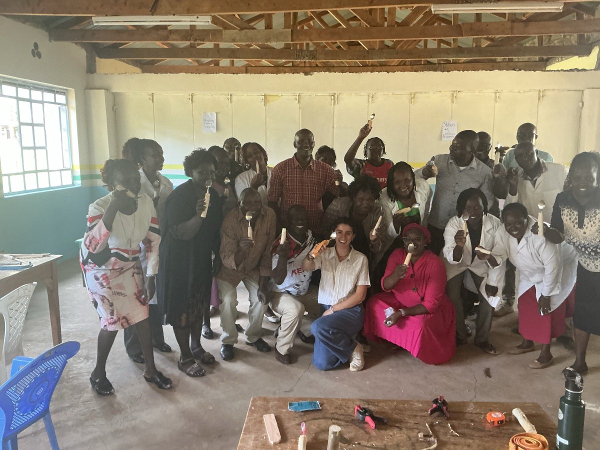 Leilani kneeling in front of the whole teaching staff in the workshop, everyone holding up the spoons with built-up wooden handles they made