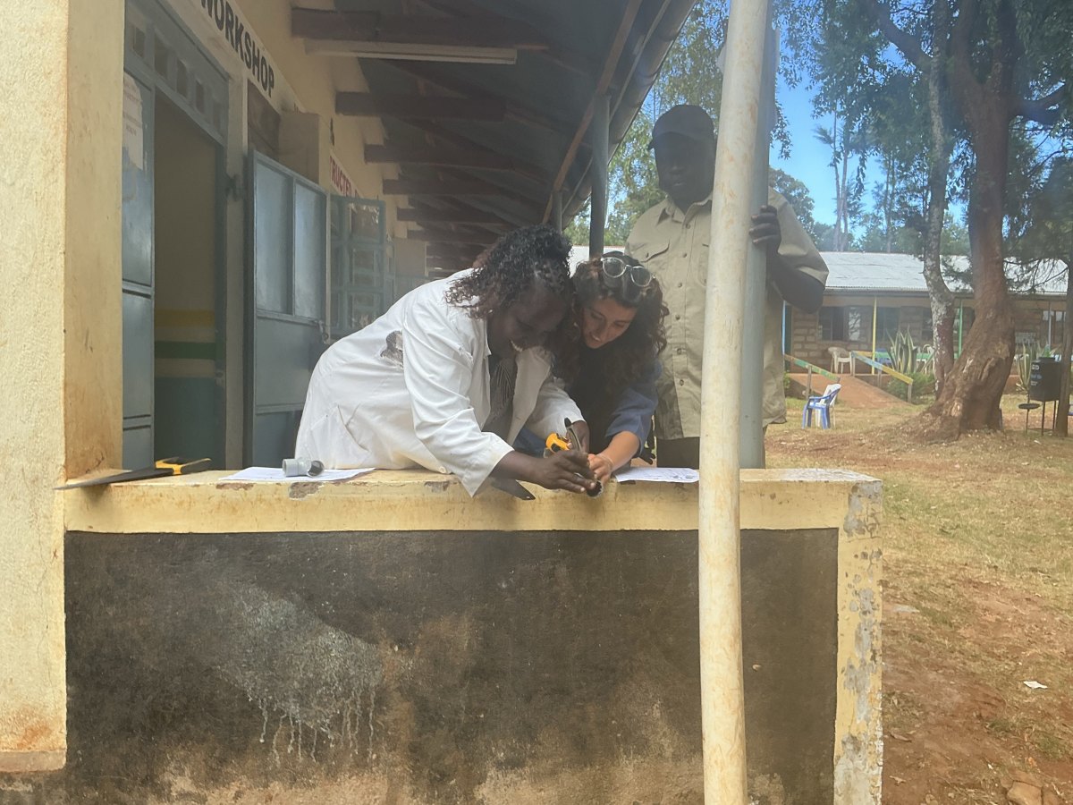 Leilani and a teacher in a white coat leaning over a low wall outside the workshop, measuring and marking PVC pipe with a tape measure and a saw beside them, while another teacher watches