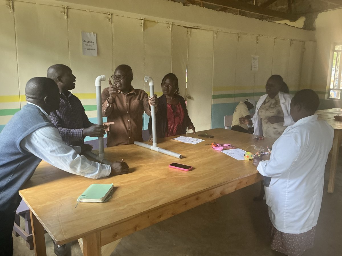 Teachers gathered around a workshop table, holding up and discussing a PVC pipe frame they are assembling