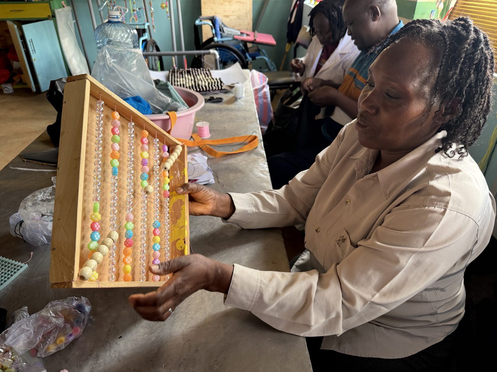 A teacher holding up a wooden frame strung with vertical rows of colorful flower-shaped and clear beads, with other teachers sewing at the table behind her