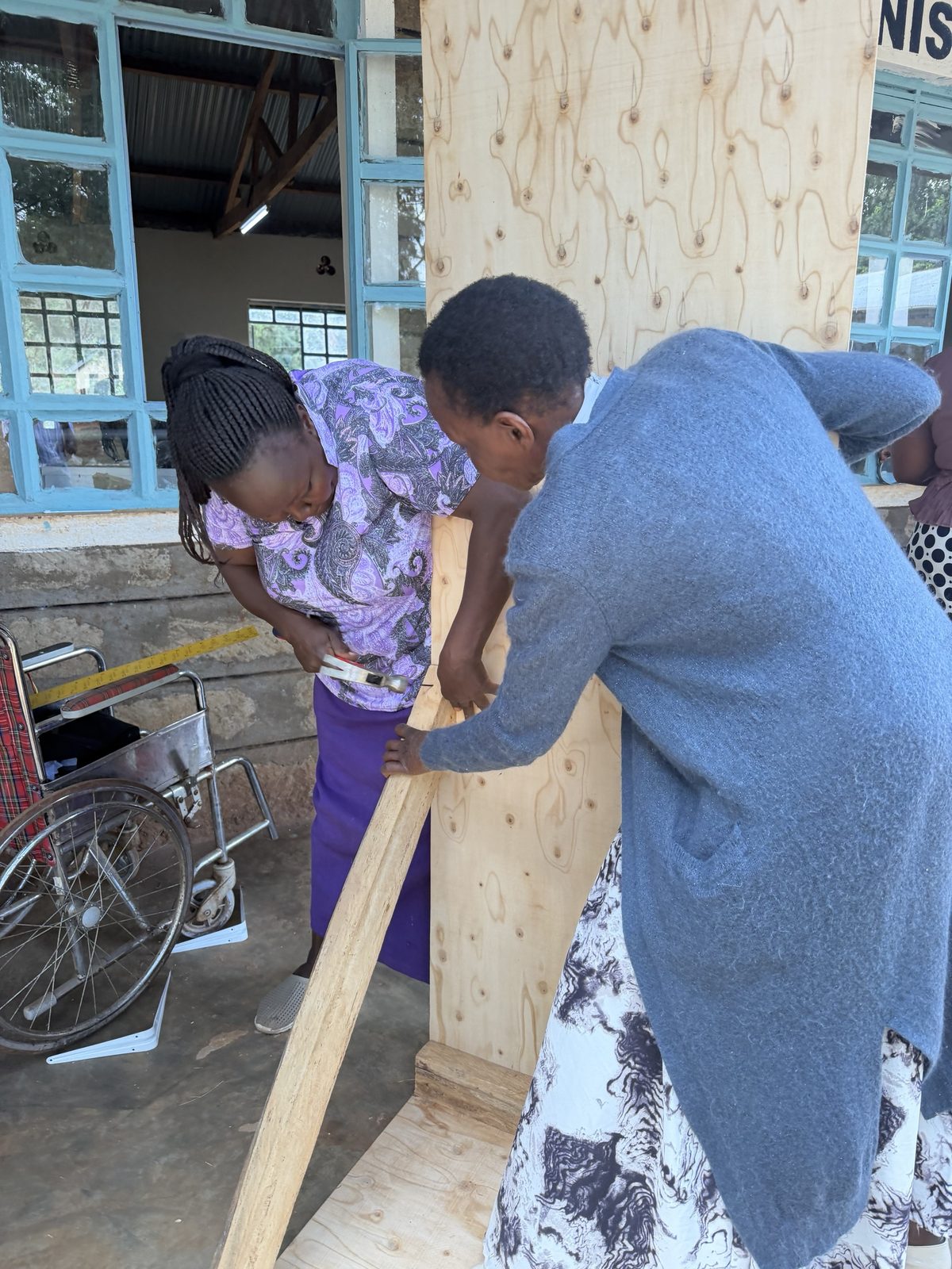 Two teachers outside the workshop hammering a wooden rail onto a tall plywood board, with a wheelchair and a measuring tape beside them