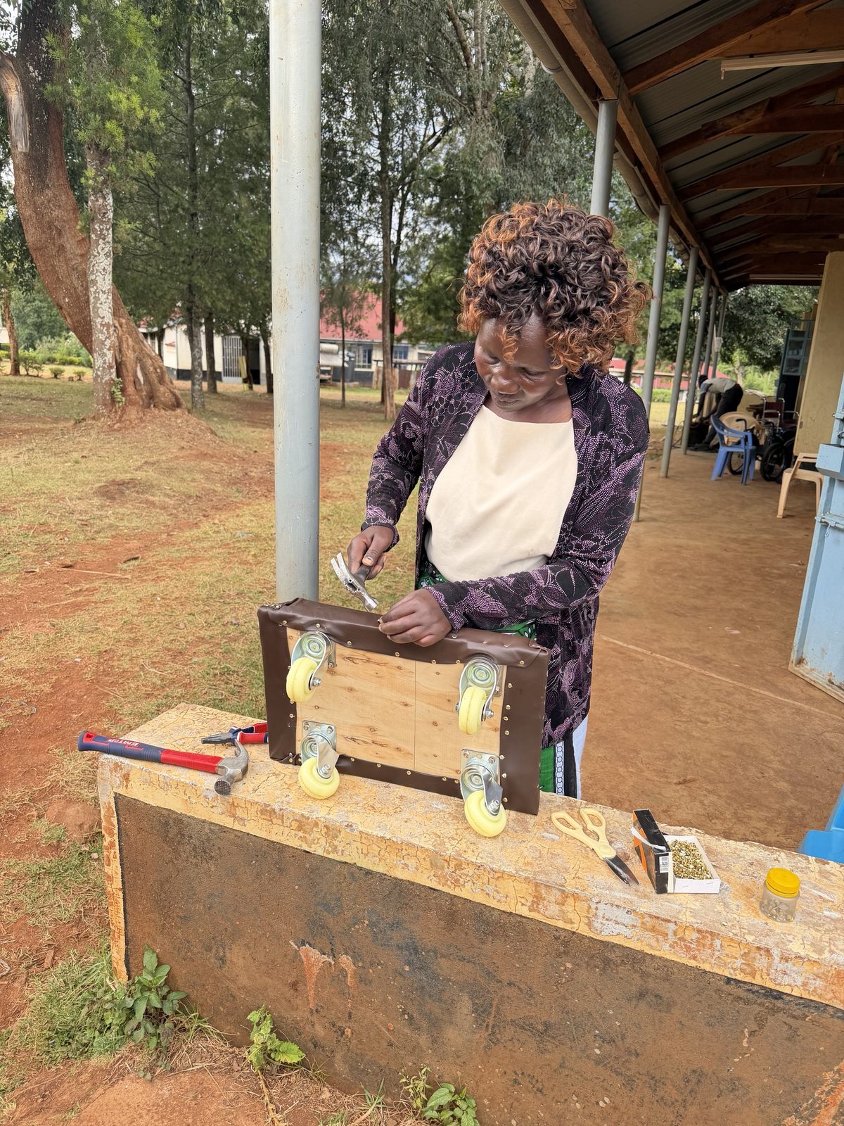 A teacher outside the workshop hammering brass tacks to fix brown vinyl around the edge of a plywood scooter board fitted with four caster wheels