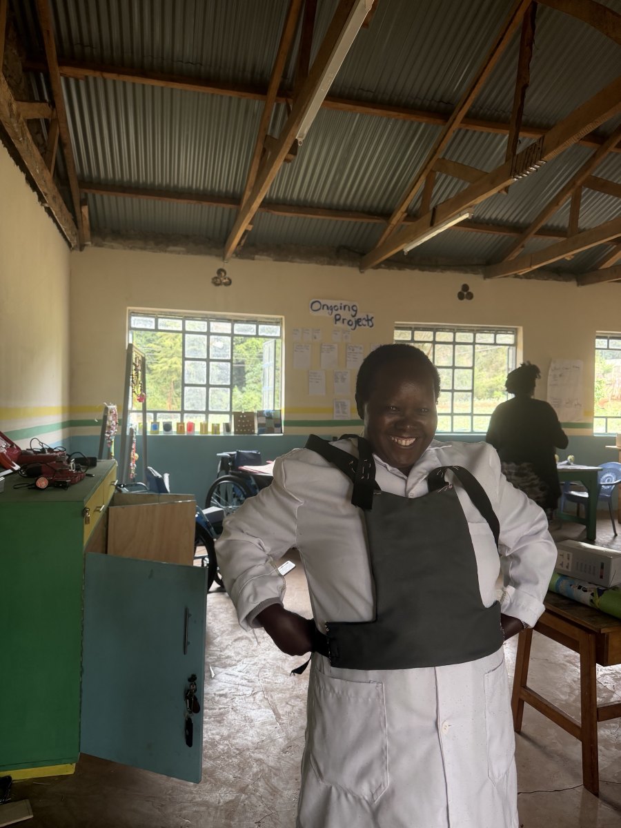 A smiling teacher in a white coat trying on a gray padded chest harness with black webbing straps in the workshop