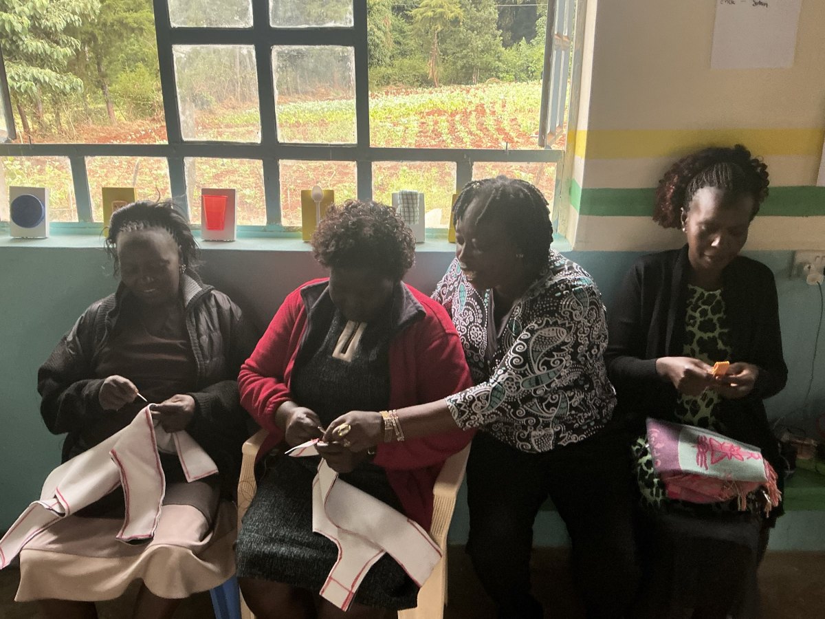 Four teachers seated by a window hand-sewing white padded harness pieces with red stitching, while one leans in to help, with finished tactile cue cards lined up on the sill