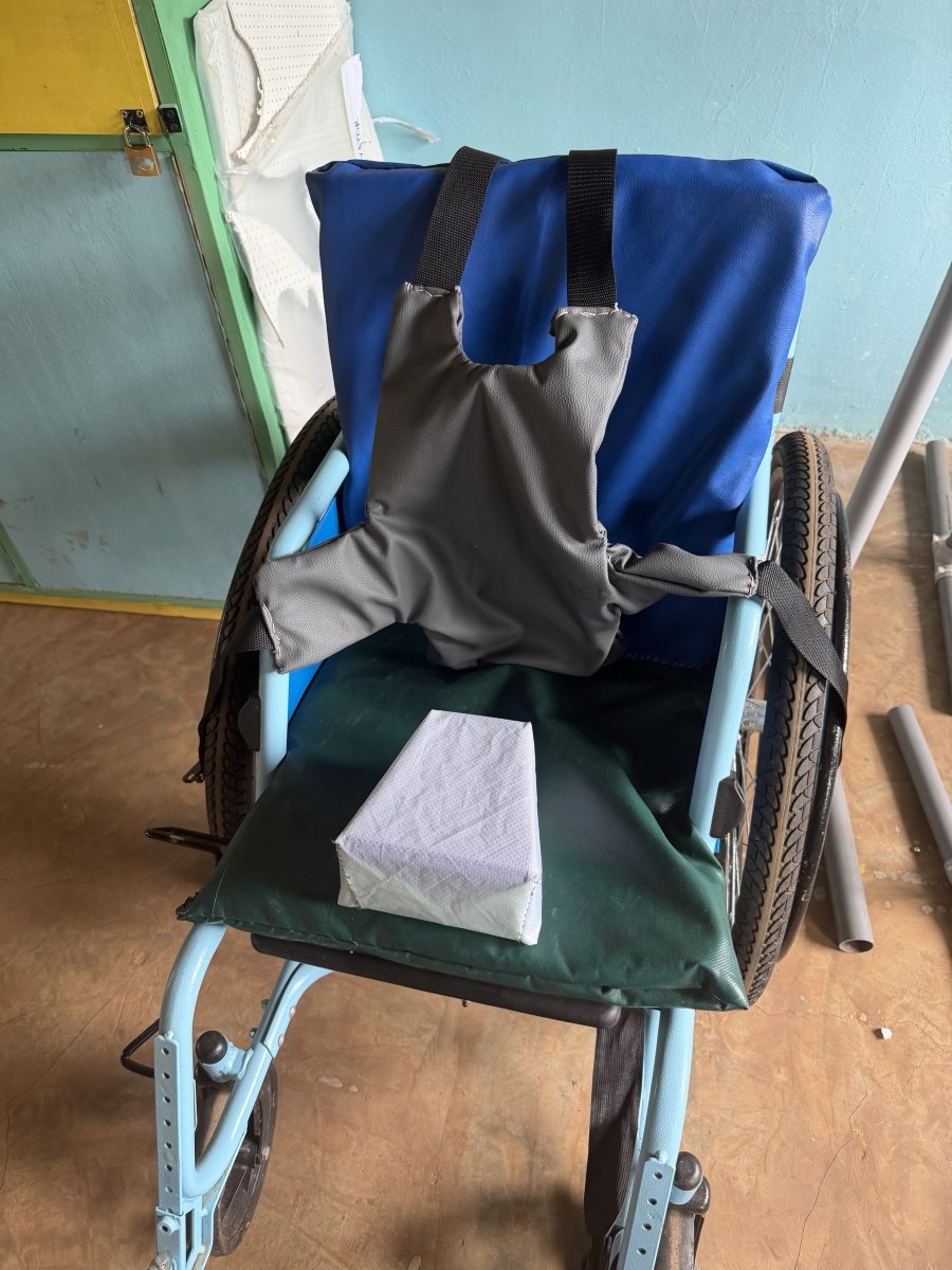 A wheelchair fitted with a gray padded chest harness on black webbing straps, a blue padded backrest, a green seat cushion and a white pommel block between the knees