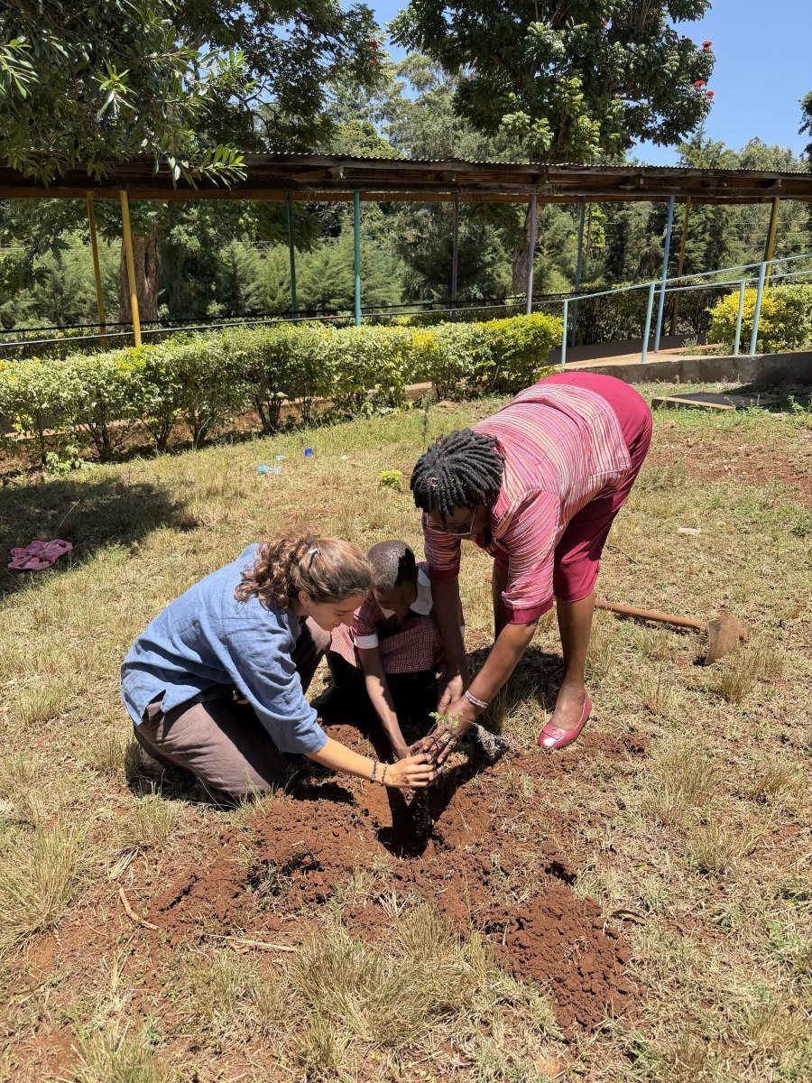 Leilani kneeling with a learner in a red checked school uniform and a teacher bending over, planting a young tree together in freshly dug red soil