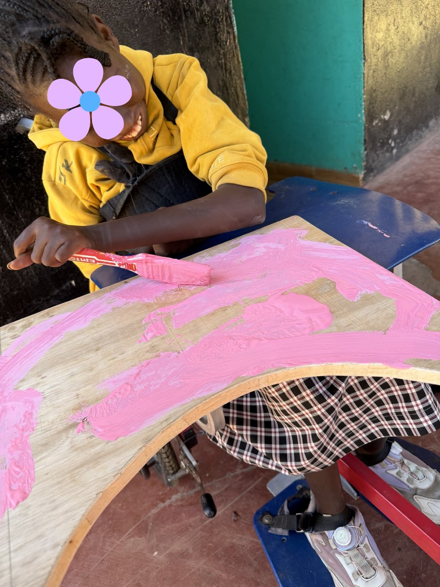 A smiling learner in a yellow hoodie, seated in her wheelchair, painting a curved wooden board pink with a brush; her eyes are covered with a flower for privacy