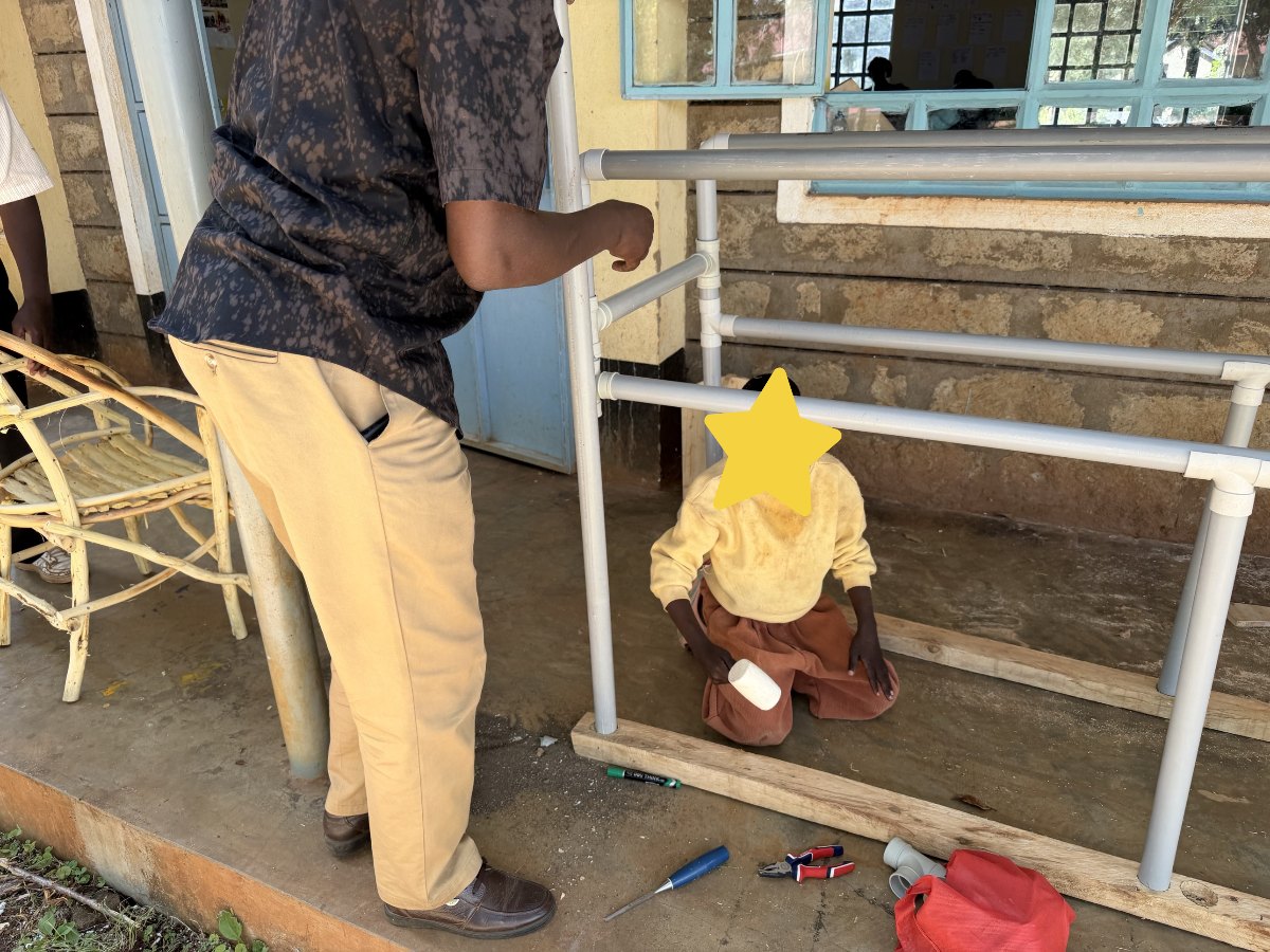 A learner in a yellow sweater kneeling on the floor inside a new set of PVC parallel bars, tapping a joint into place with a white mallet, while a teacher stands beside her; her face is covered with a star for privacy