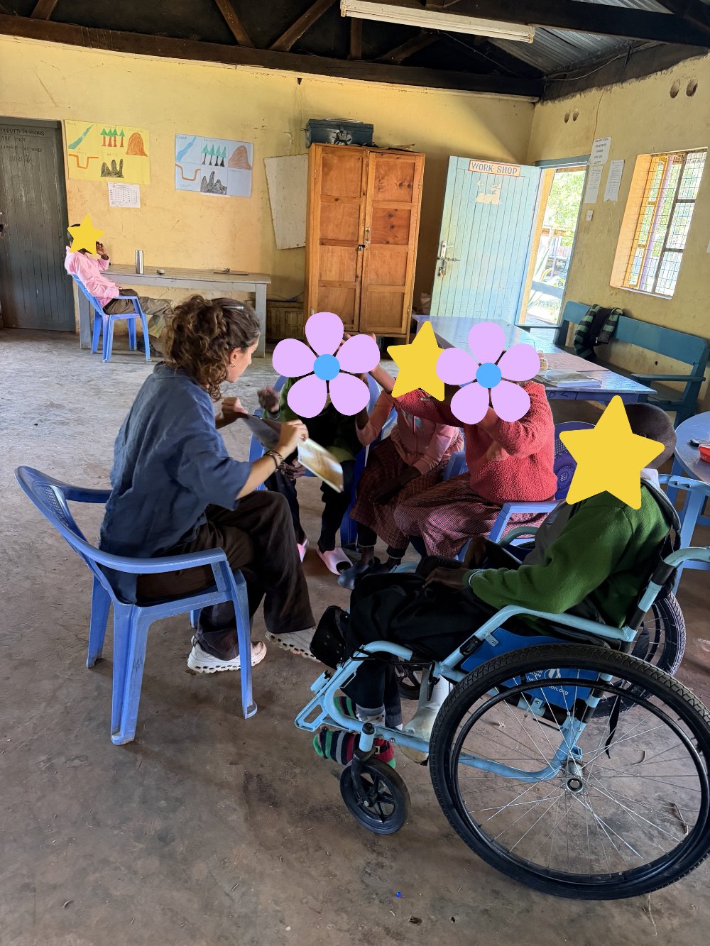 Leilani reading aloud from a book to a circle of learners, one in a wheelchair, who are reaching out and signing excitedly; their faces are covered with flowers and stars for privacy