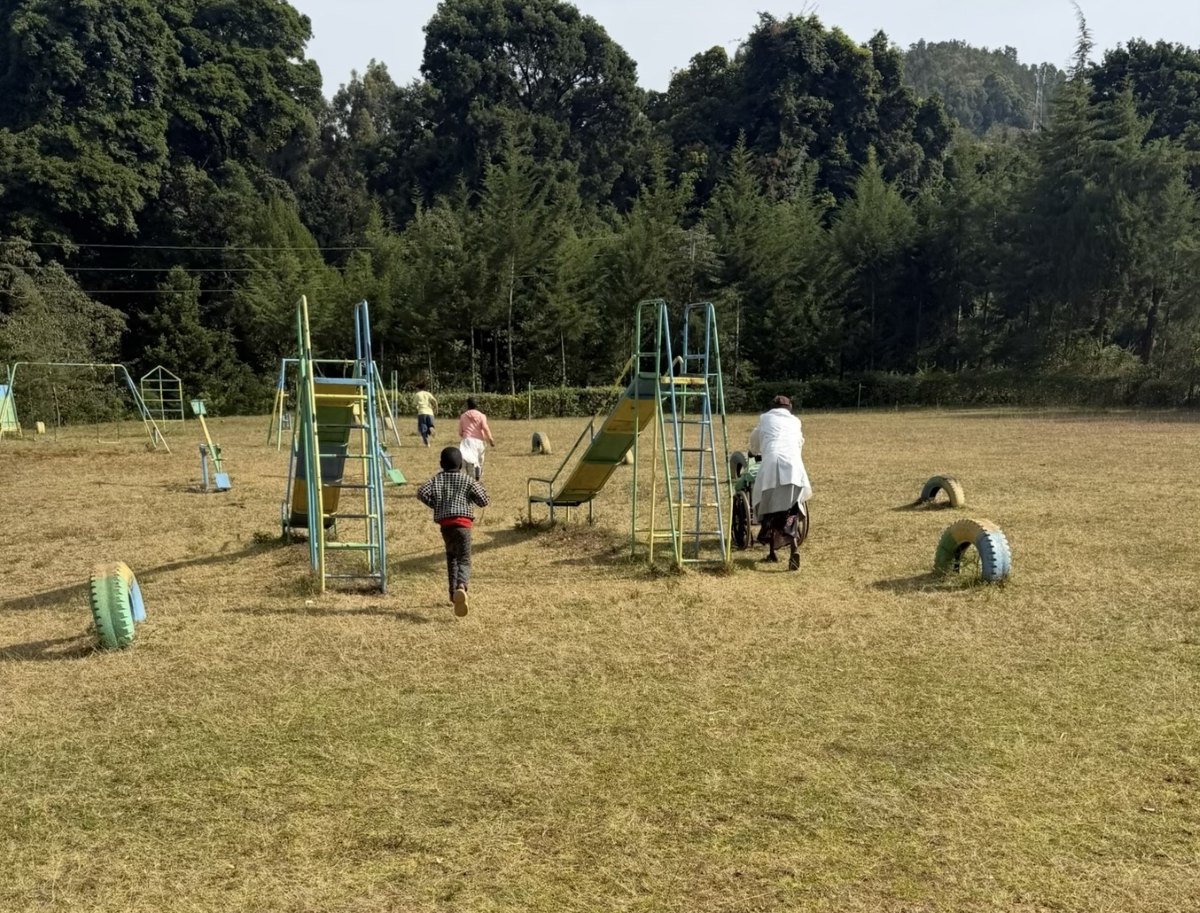 Learners running across the school field toward the playground, seen from behind, with a teacher pushing a wheelchair