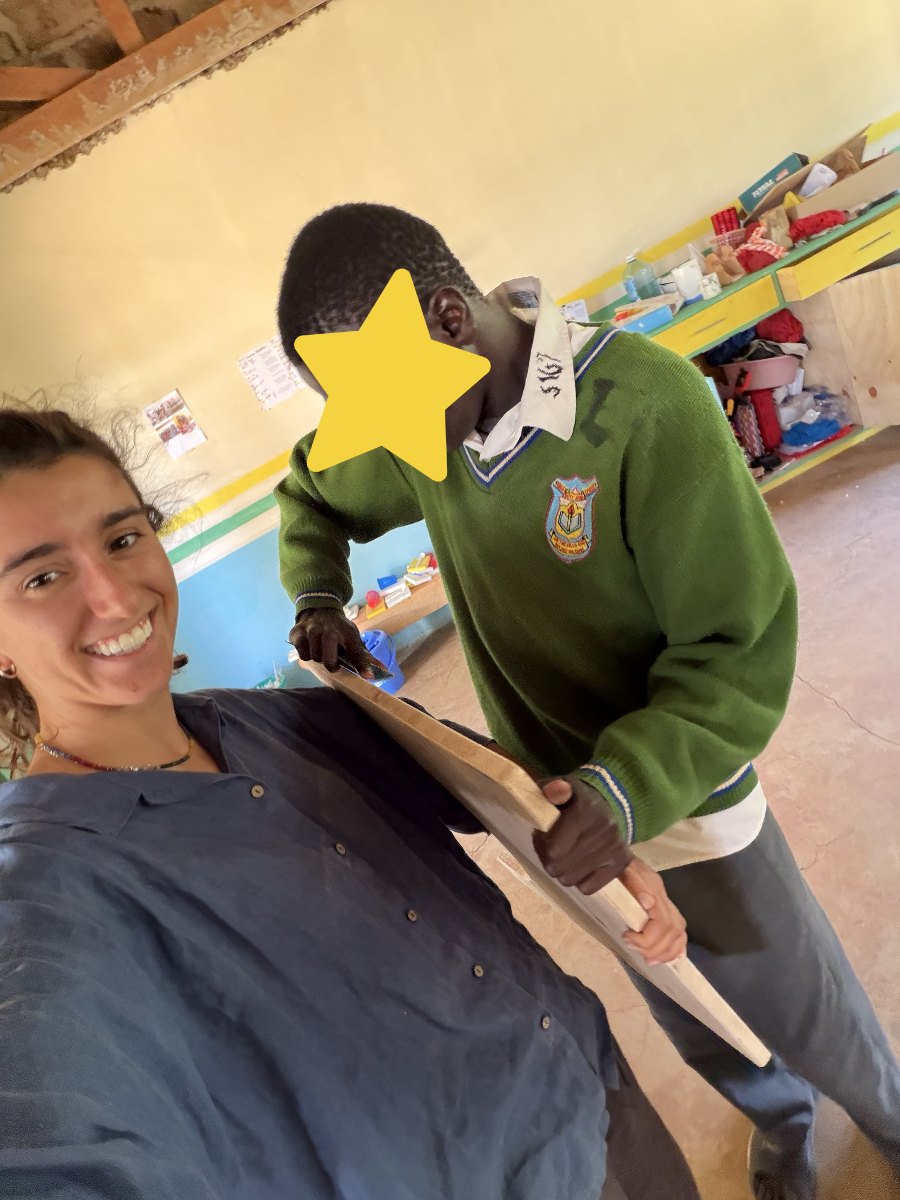 Leilani smiling in a selfie beside a learner in a green school sweater who is sanding the edge of a wooden board; his face is covered with a star for privacy