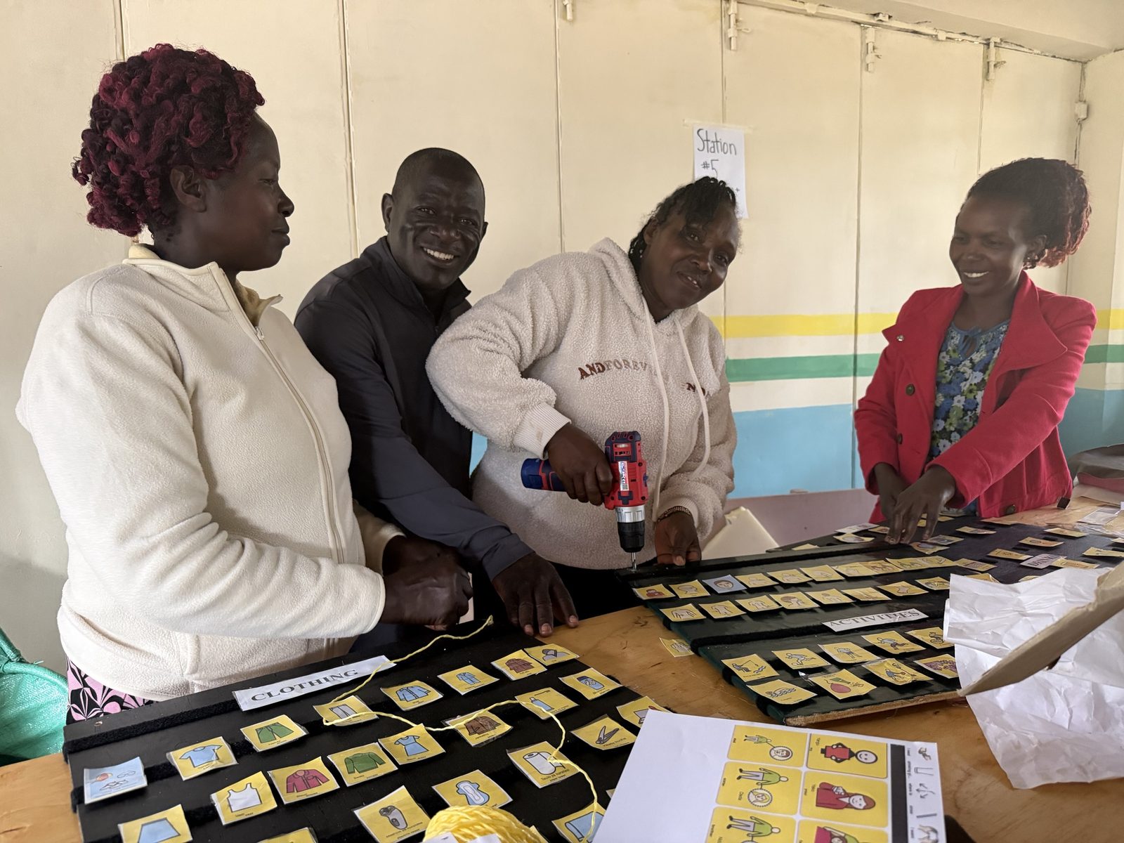 Four teachers smiling around a workbench as one drills a strip onto a black communication board, with rows of yellow picture symbol cards sorted under Clothing and Activities headings