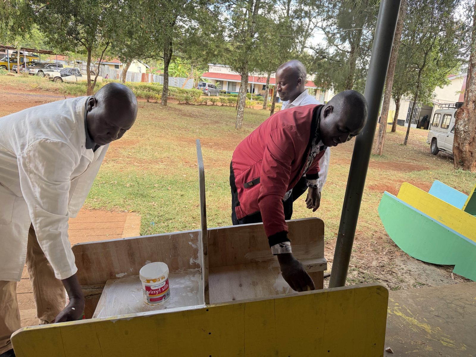 Three teachers outside the workshop priming a plywood two-person rocker with white paint, with a finished rocking chair painted green, yellow and blue on the grass behind them