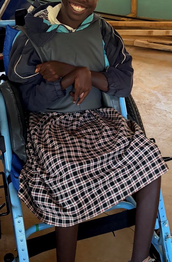 A smiling learner seated in a wheelchair wearing a gray padded chest harness