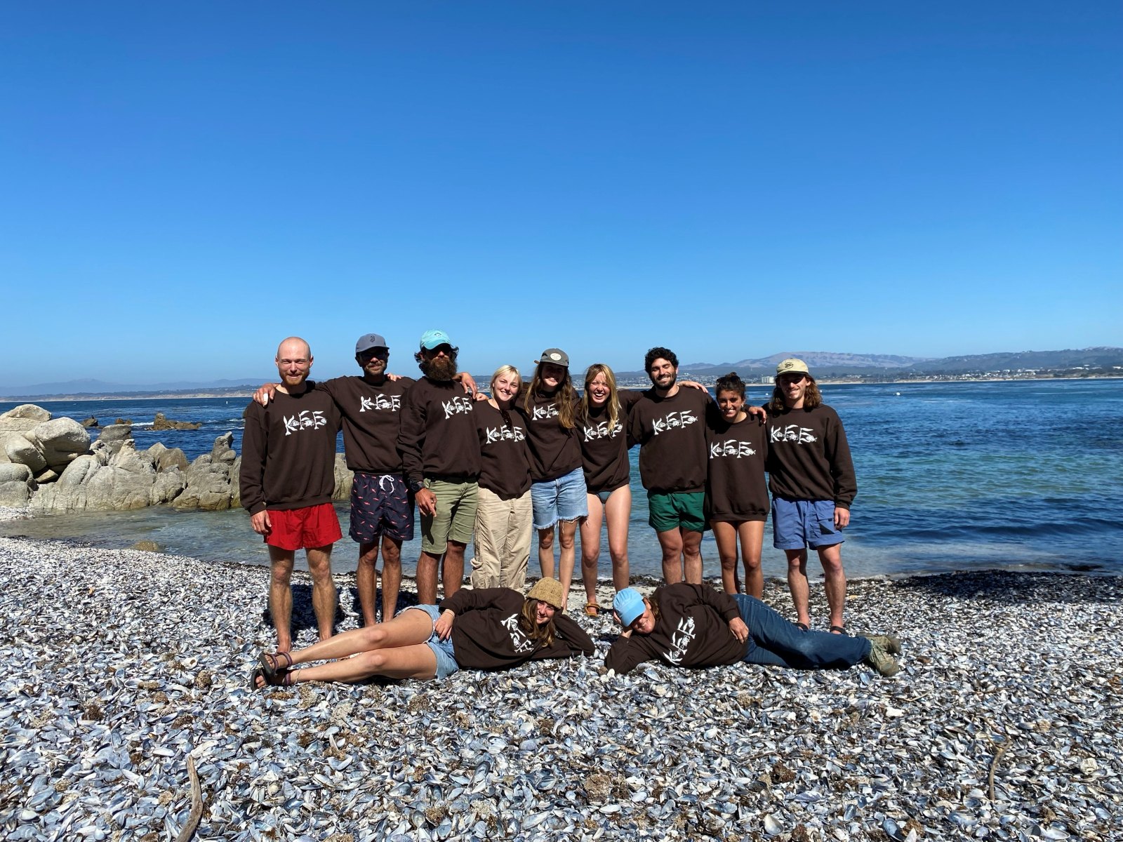 The Kelp Forest Ecology class in matching brown KFE sweatshirts on a shell-covered beach at Hopkins Marine Station, with Monterey Bay behind them