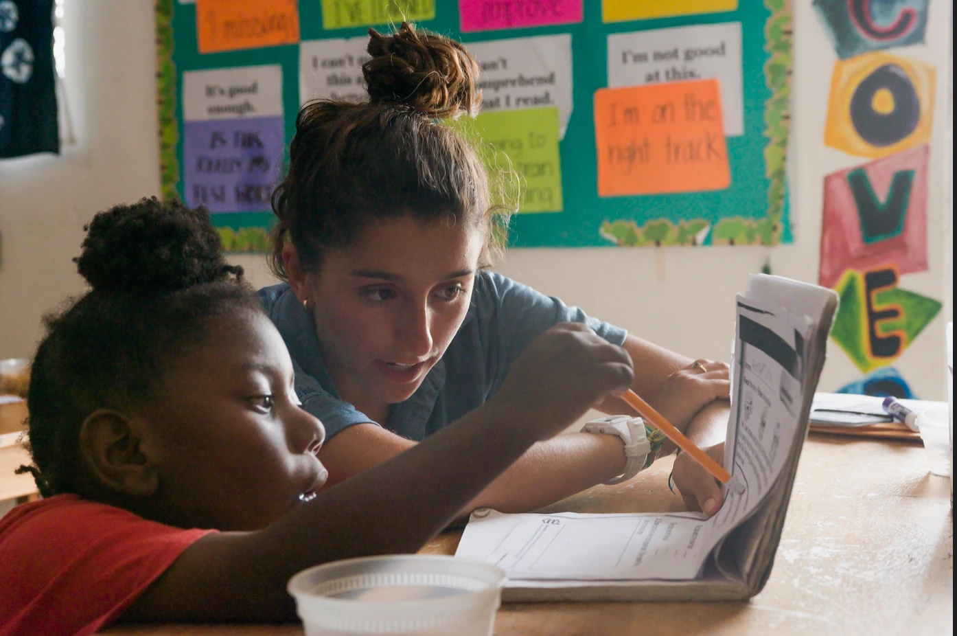 An Island School student leaning in to help a young reader work through a book at a classroom table