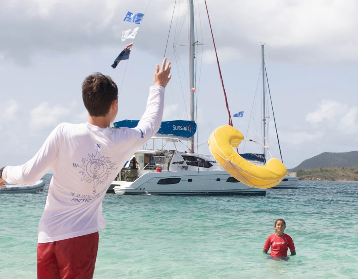 A student throwing a yellow ring buoy to a swimmer during a water rescue drill, with catamarans behind