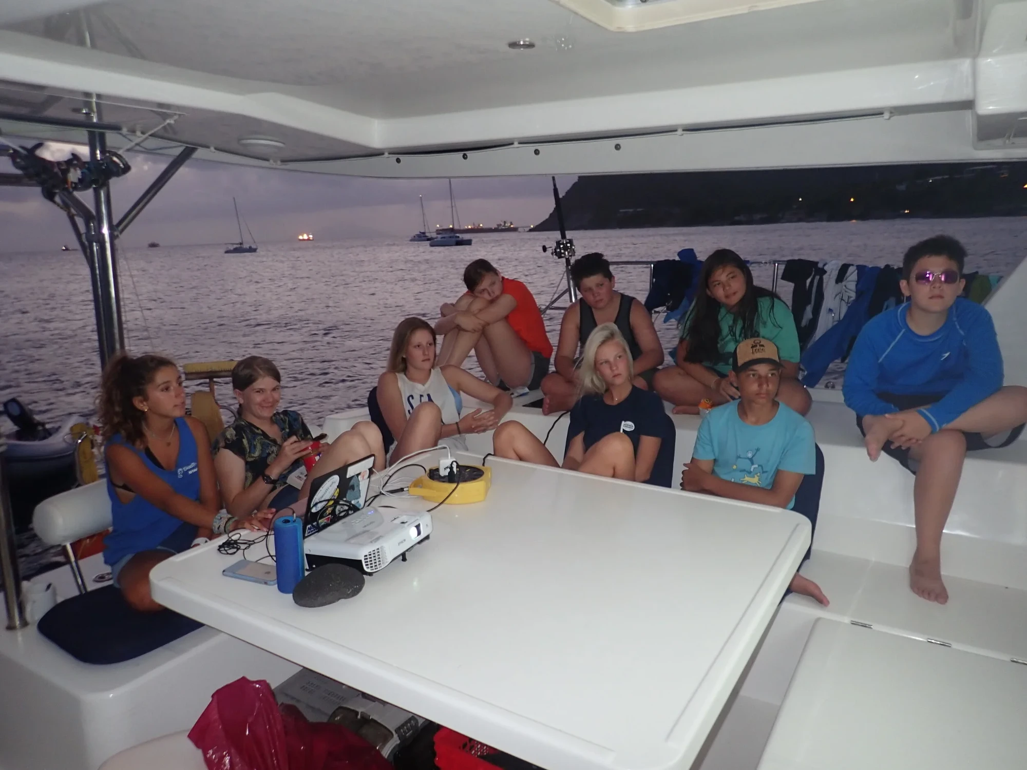 Students gathered on a catamaran deck at dusk watching a projector presentation